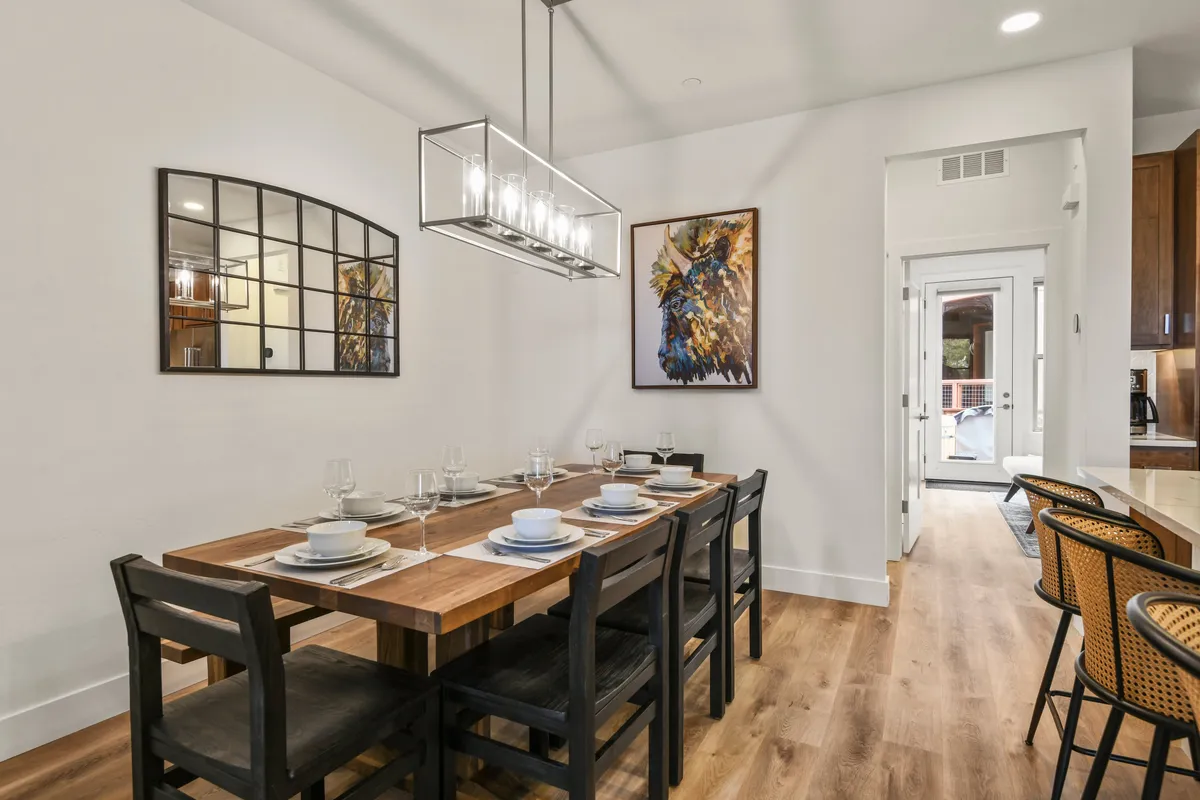 A beautifully set dining table under a shimmering chandelier, complemented by wall art and a mirror that enhances the sense of space. The arrangement and decor invite guests to sit down for a meal, play a board game, or celebrate special occasions together.