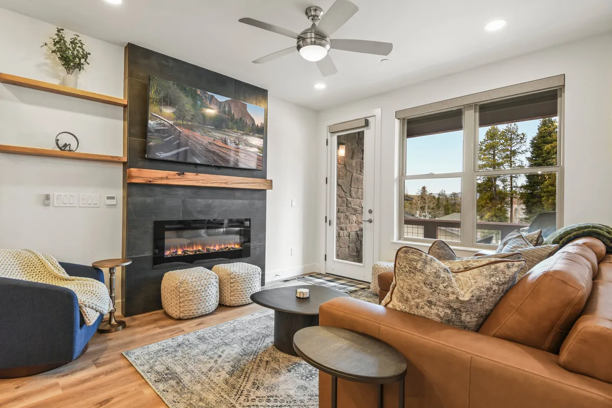An inviting living room featuring a prominent fireplace with flames glowing, a large TV mounted above or nearby, and deeply cushioned, comfortable seating arranged to foster conversation and relaxation. Through the large windows, natural light pours in, and an outdoor vista is visible, hinting at the Colorado scenery beyond.