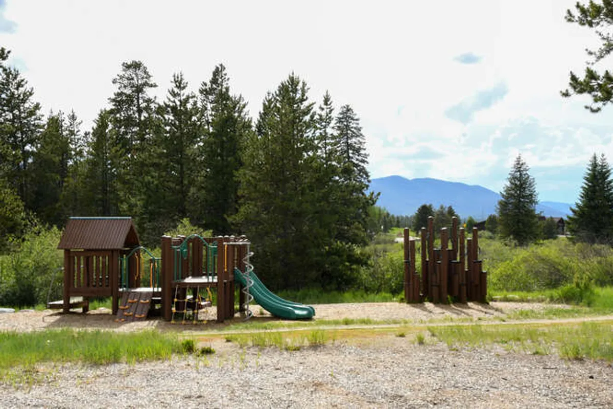 A cheerful playground scene with colorful slides and equipment surrounded by leafy trees. The area is clearly designed for families, with children playing and parents gathering nearby. Nature trails can be seen leading away from the playground, inviting further exploration.