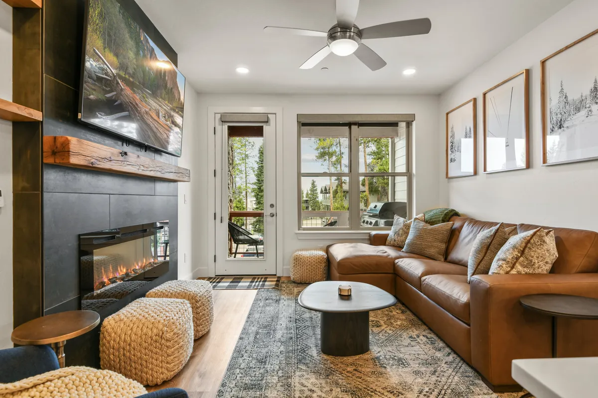 The image showcases a warm, inviting living room with a plush leather sofa centered around a crackling fireplace, a modern television, stylish coffee table, and tasteful artwork adorning the walls. Large windows fill the space with natural light and offer a glimpse of mountain sunsets, creating a perfect setting for relaxation and connection.