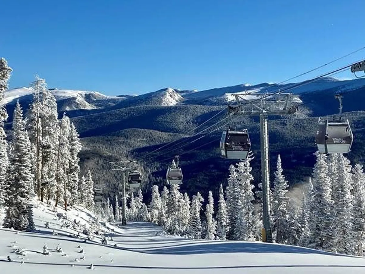 A scenic exterior view of a gondola lift rising above snow-covered trees and mountain slopes, set against a clear blue sky. The image evokes the thrill of skiing and snowboarding, while also highlighting the breathtaking natural beauty of the area.