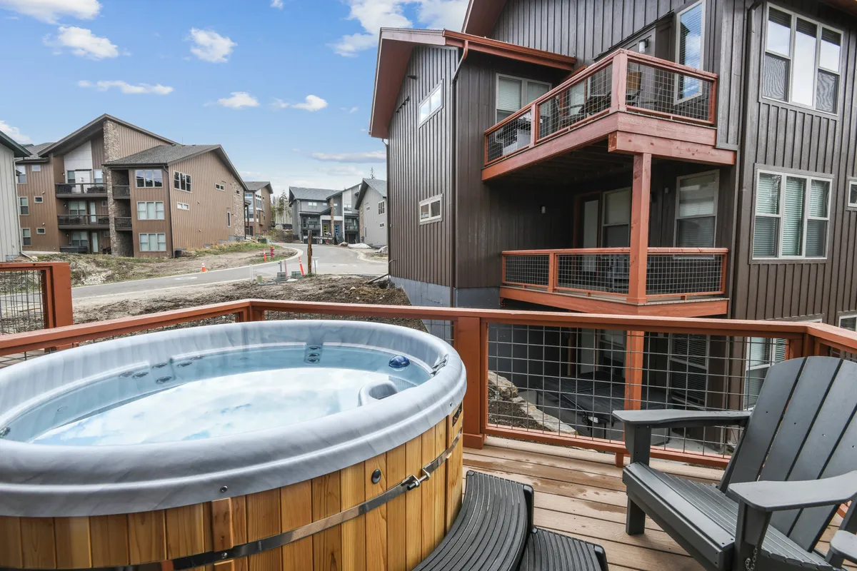 This image showcases a spacious deck with a hot tub as its focal point, surrounded by adjacent buildings. The scene captures people relaxing in the bubbling hot tub and others socializing on the deck, all set against a serene backdrop. The overall mood is inviting and leisurely, emphasizing comfort and opportunities to unwind after outdoor adventures.