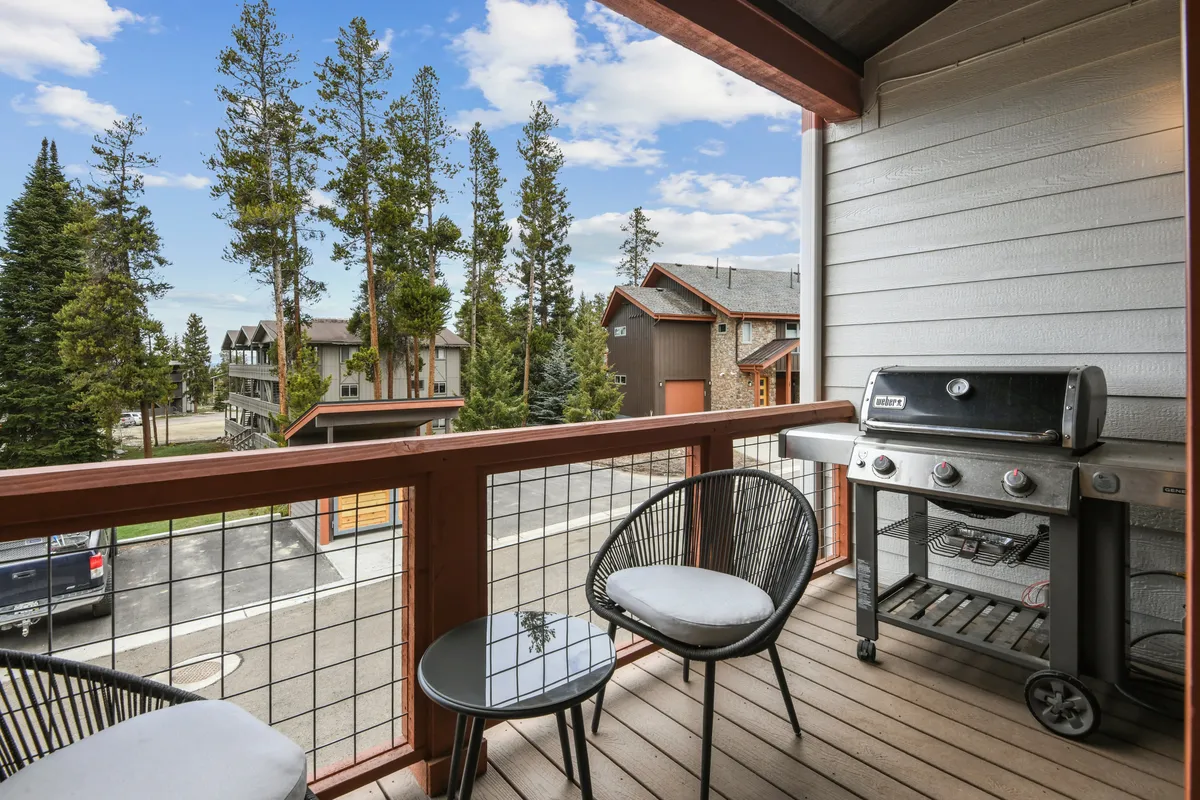 This image features a balcony or patio area with comfortable seating and a gas grill, surrounded by lush green trees. The setting suggests a perfect spot for outdoor dining, relaxing with a drink, or grilling up a mountain BBQ while enjoying views of nature.