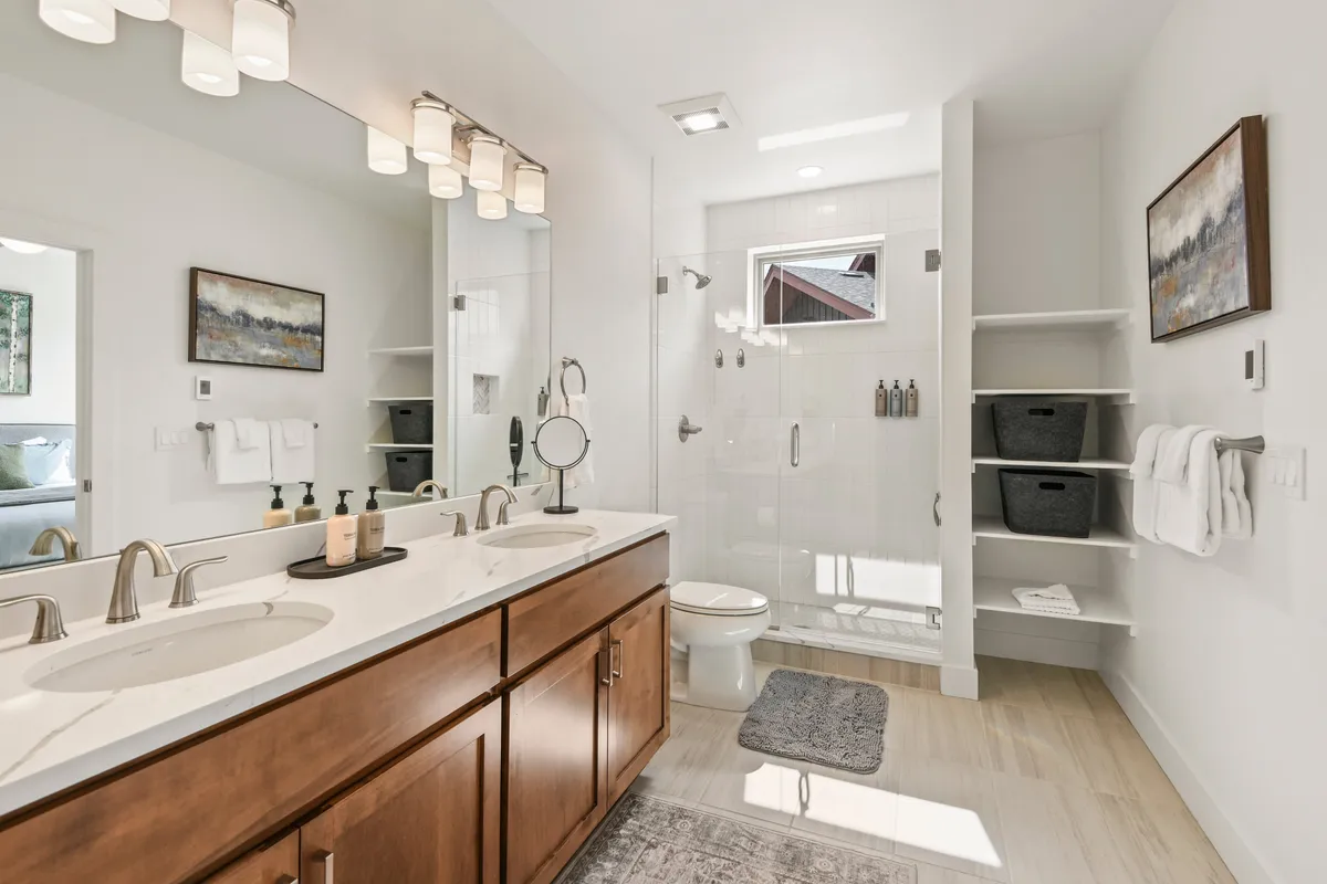 A serene en-suite bathroom with a double sink vanity, elegant fixtures, glass shower enclosure, and tasteful artwork. Organized open shelving and calming tones make the space feel luxurious and spa-like.