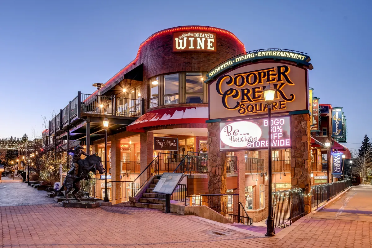 This exterior scene shows the lively Cooper Creek Square with its prominent signage, wine shop, outdoor seating, and entrance stairs. The festive mood and visible gathering spaces hint at dining, entertainment, and après-adventure socializing.